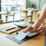 Expert comptable restauration travaillant sur ordinateur portable et calculatrice dans un restaurant moderne, gestion des obligations fiscales 2025, table en bois clair avec documents financiers, ambiance professionnelle chaleureuse.