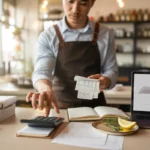 conseils comptables restaurateurs débutants : vue oblique au-dessus de l’épaule des mains d’un jeune restaurateur triant des reçus et utilisant une calculatrice, carnet ouvert et ordinateur portable aux graphiques abstraits, herbes et citron sur comptoir en bois clair; lumière naturelle, bokeh