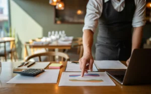 Comment faire un business plan efficace pour un restaurant: mains d’un restaurateur en veste de cuisine pointant un graphique sur table en bois, laptop aux graphiques colorés sans texte, calculatrice, feuilles et plan de salle, post-it, herbes; lumière dorée, arrière-plan de restaurant flou.