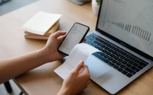 Comment passer à la digitalisation de sa comptabilité: main scannant un reçu vierge avec smartphone sur bureau moderne, laptop aux graphiques abstraits, petite pile de papiers, transition papier vers numérique.