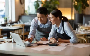 Accompagnement comptable dans un restaurant moderne : un comptable et un restaurateur collaborent côte à côte devant un ordinateur portable affichant des graphiques flous, avec classeur, calculatrice et documents sur une table en bois clair, dans une ambiance chaleureuse et professionnelle.