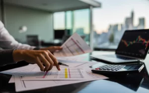 Vignette photo réaliste d’un bureau moderne montrant les mains d’un comptable en plein audit comptable, examinant des bilans imprimés et des graphiques de risques sur ordinateur dans un open space lumineux, contexte entreprise 2025.