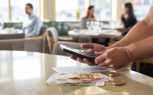 Comptoir de restaurant moderne en bois clair avec mains d’un restaurateur utilisant une calculatrice et gérant une addition détaillée, pièces et billets d’euro visibles, ambiance chaleureuse ; illustration du concept tva en restauration.